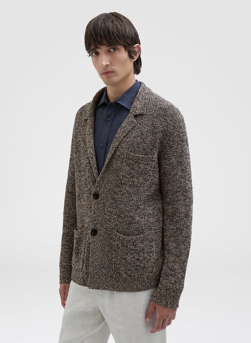 Man in a brown knit blazer and blue shirt, posing indoors against a white background. Fashionable menswear, studio shot.