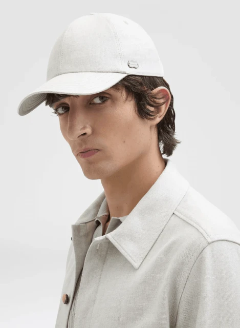 Close-up shot of a young adult man wearing a light gray baseball cap and matching shirt against a white background in a studio setting.