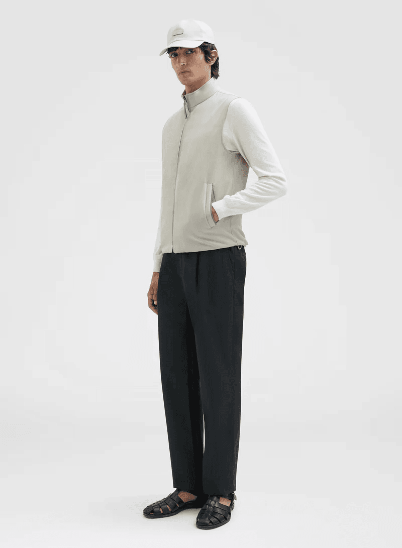 A full-length studio shot captures a slender man in a light-colored vest, long-sleeved shirt, and dark trousers, complemented by a baseball cap.