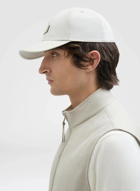 Profile view of a young adult man wearing a light-colored cap and vest against a bright white backdrop in a studio setting.