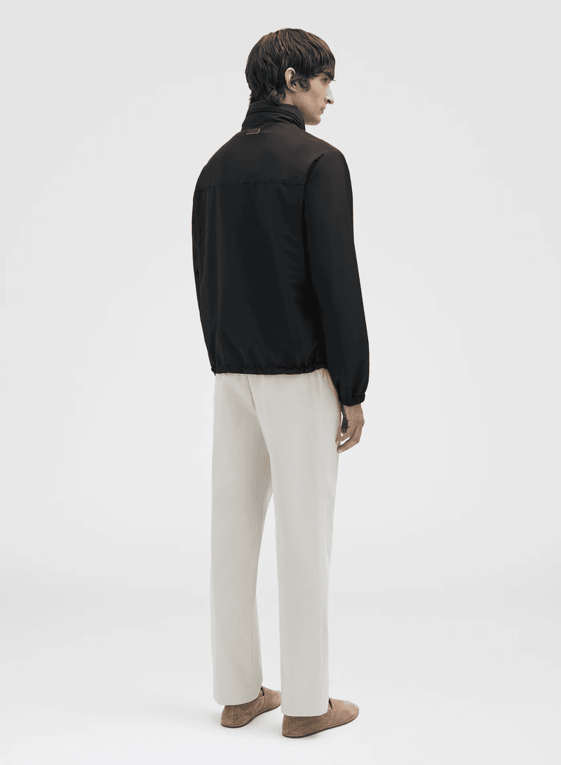 Back view of a male model wearing a black jacket and light beige pants against a white background in a fashion studio shot.