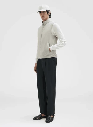 Male model wearing a cream zip vest over a long-sleeve white top, dark trousers, cap, and black sandals against a plain background