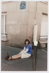 A woman seated on a Paris street pavement wearing a blue striped top, cream skirt, and lace-up brown block heels.