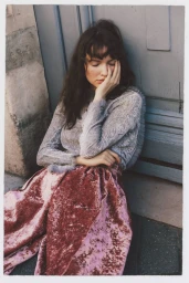 Woman with dark wavy hair wearing a sheer lavender lace top and crushed velvet skirt leaning against a blue wooden door.