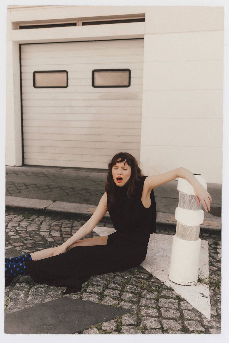 A woman in a black sleeveless dress and patterned blue socks sits on a cobblestone street leaning against a white bollard.