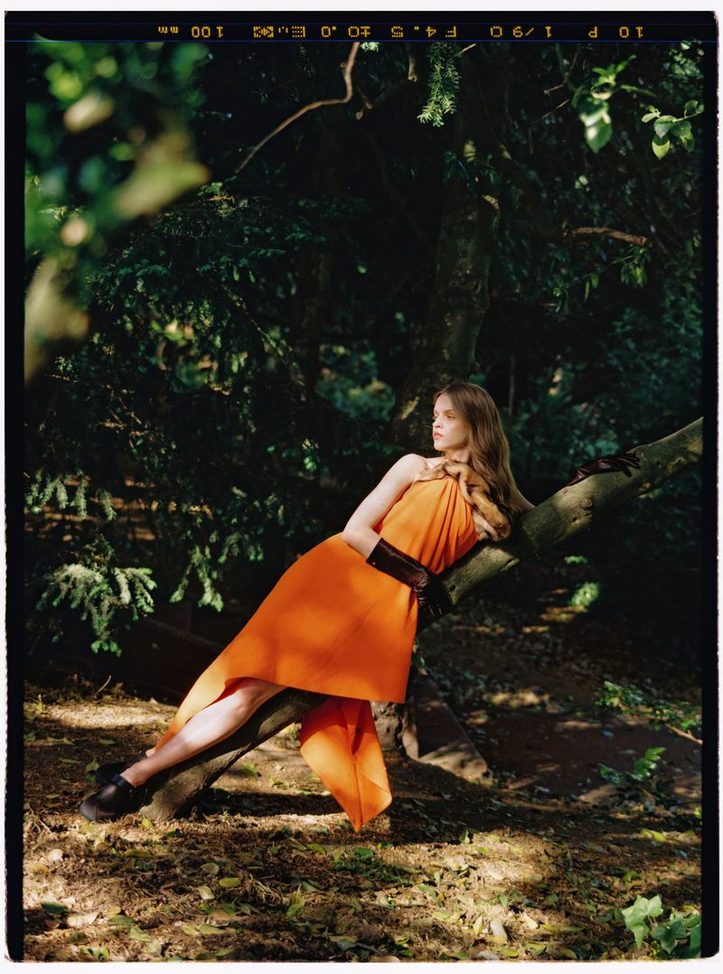 Model Batura Aliaksandra in an orange dress and dark gloves reclining on a tree branch in a woodland setting.