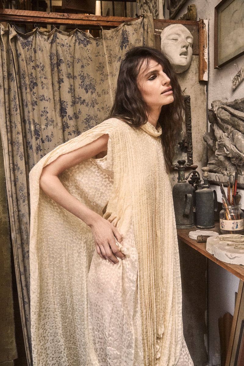 A woman with long brown hair wearing a flowing cream pleated cape dress, posed in a cluttered artist studio with plaster sculptures.