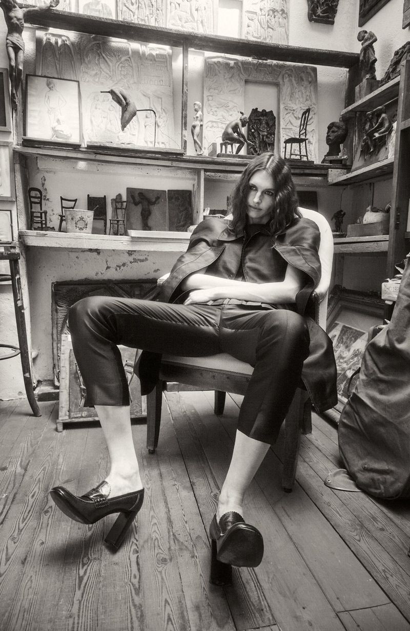 Model Angelika Baranska seated in an armchair in an art studio, wearing dark tailored trousers and high-heeled loafers.