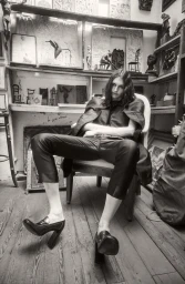 Model Angelika Baranska seated in an armchair in an art studio, wearing dark tailored trousers and high-heeled loafers.