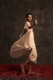 Model Angelika Baranska posing in a flowing cream dress and lace-up sandals against a textured dark background.