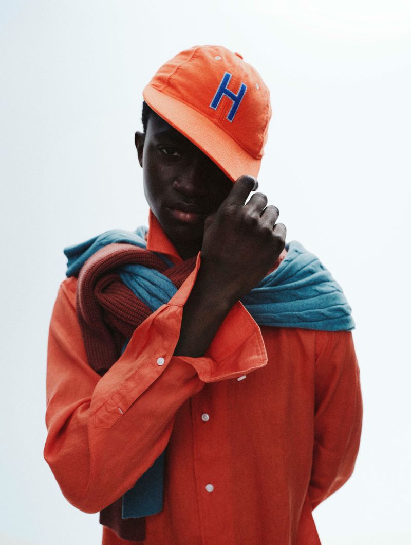 Model wearing an orange baseball cap with a blue H and an orange button-down shirt with layered knit sweaters draped over his shoulders.