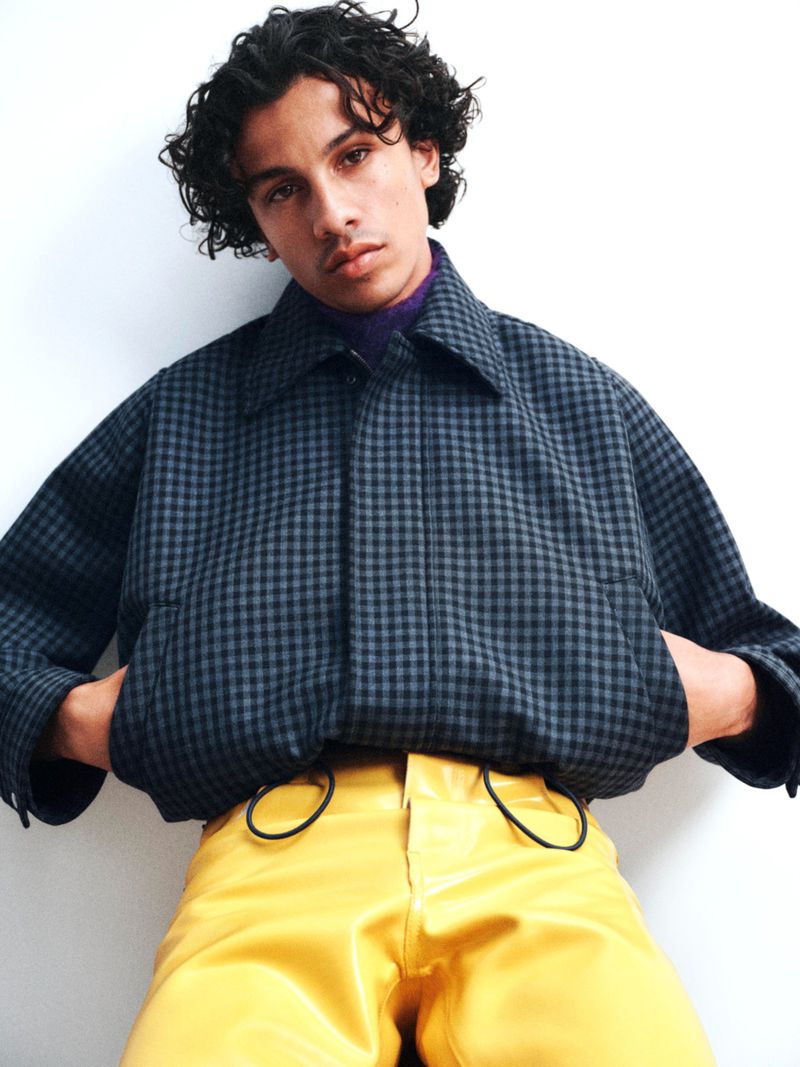 Model Ned Crapper wearing a dark checkered collared jacket and yellow trousers against a white studio background.