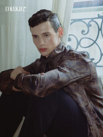 Model Paolo Borle sits near a sheer curtain window wearing a brown paisley patterned shirt and black trousers.