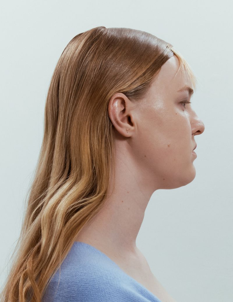 Profile portrait of a woman with styled honey blonde hair wearing a light blue top against a pale grey seamless background.
