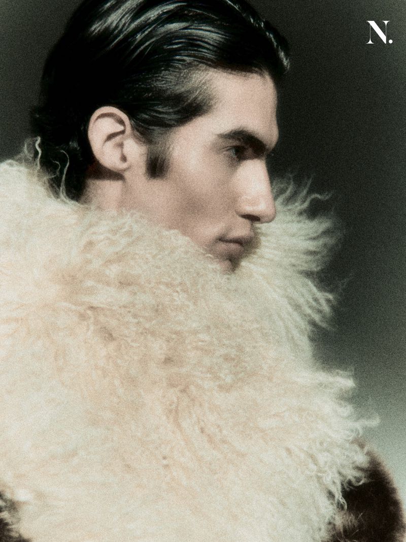 Side profile of a man with slicked-back dark hair wearing a voluminous cream shearling fur collar against a dark backdrop.