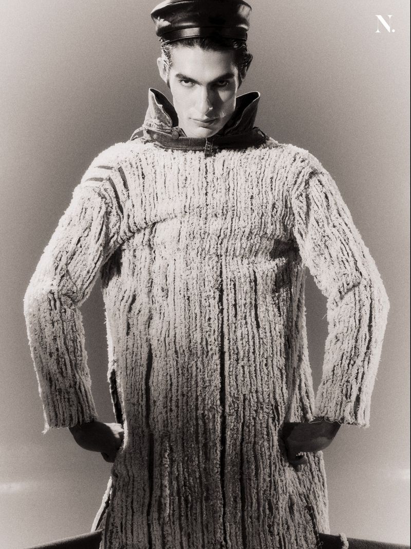 Male model in a heavily textured high-collar coat and black leather peaked cap posing with hands on hips against a studio backdrop.