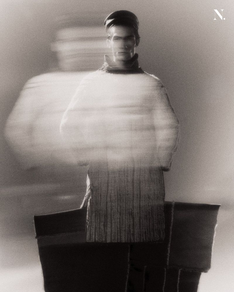 Monochrome fashion portrait of a man wearing a high-neck knit tunic and cap with motion blur effect.