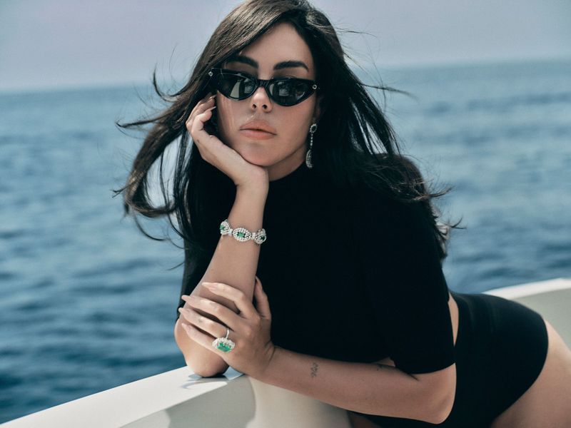 Georgina Rodriguez in black sunglasses and a high-neck swimsuit with emerald jewellery leaning on a boat railing over the ocean.