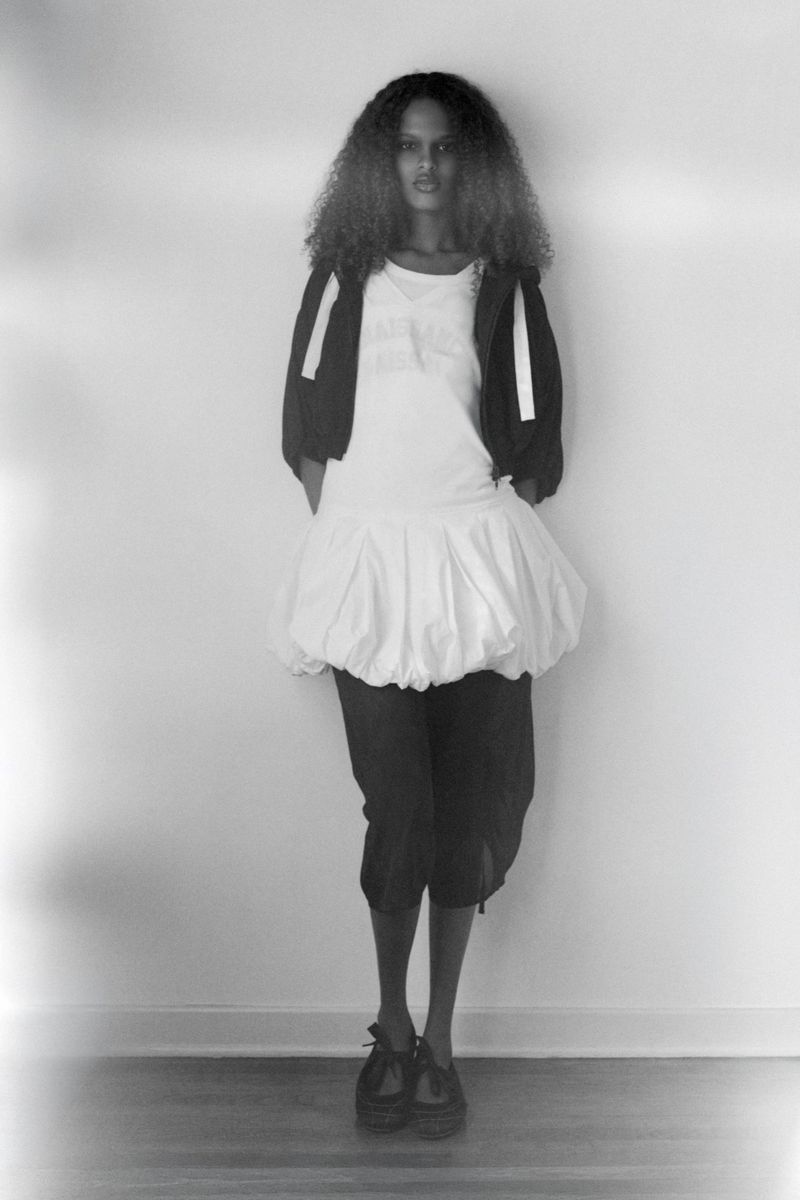Model with long curly hair wears a white bubble hem dress and dark bolero jacket standing against a plain white wall.