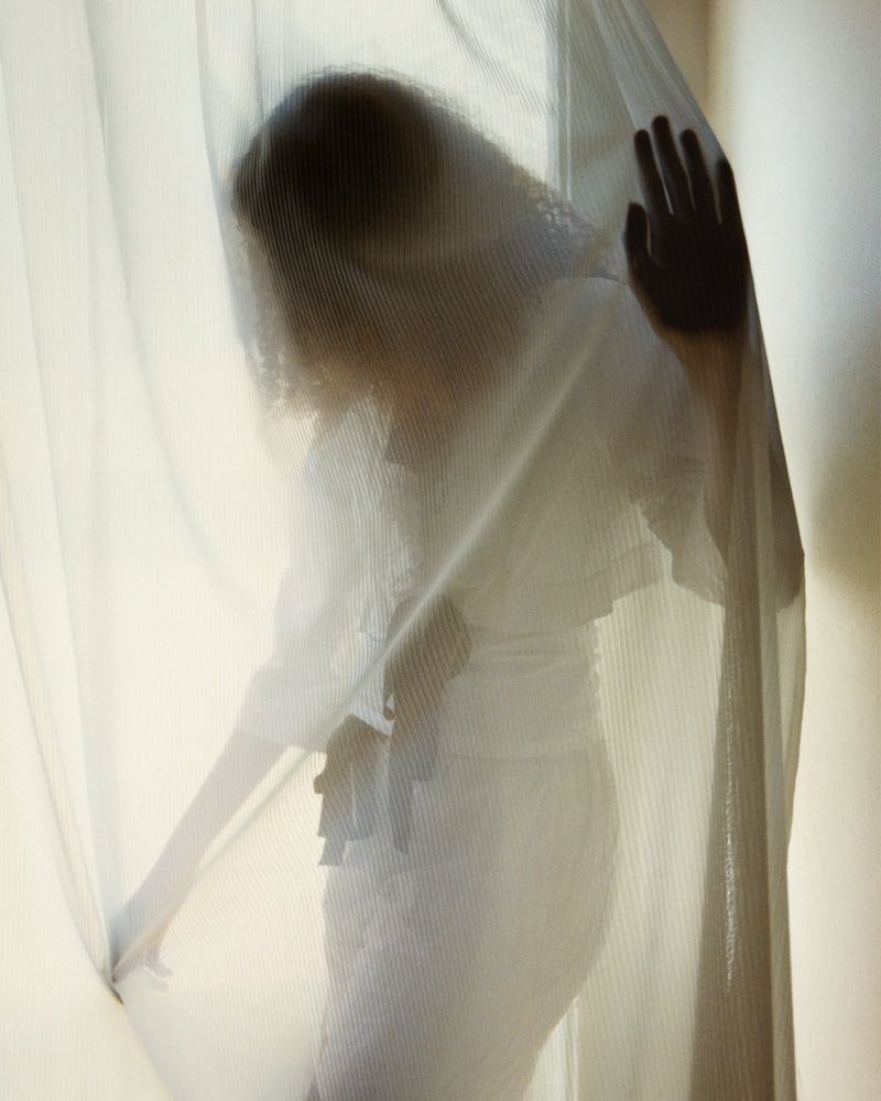 Model Yusra Saeed posing behind a translucent sheer white curtain wearing an all-white outfit.