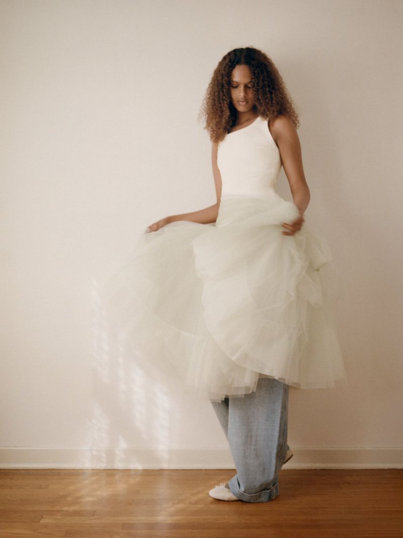 Model in a white sleeveless top and voluminous tulle dress layered over wide-leg denim jeans standing on a wooden floor.