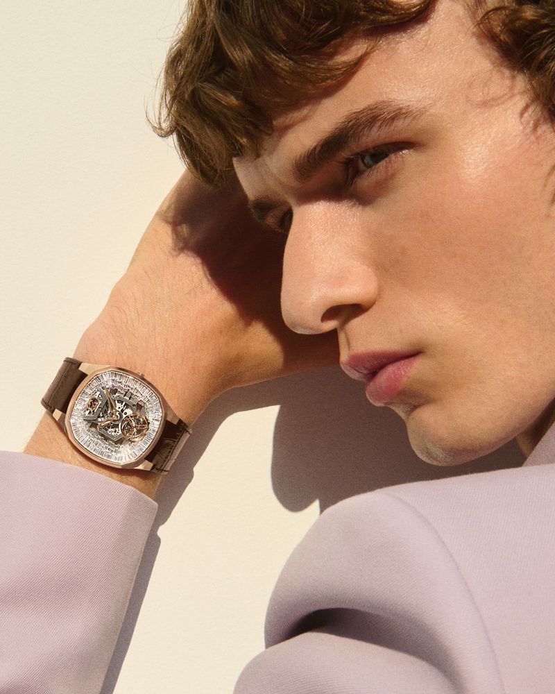 Close-up of a fashionable adult man wearing a brown leather wrist watch and a lavender blazer against an off-white background.