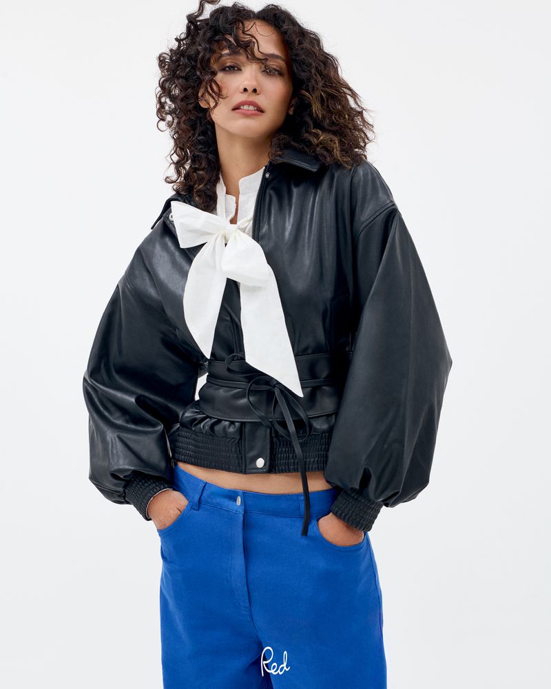 Model Cora Emmanuel in a black cinched leather jacket, white bow blouse, and blue trousers against a white background.