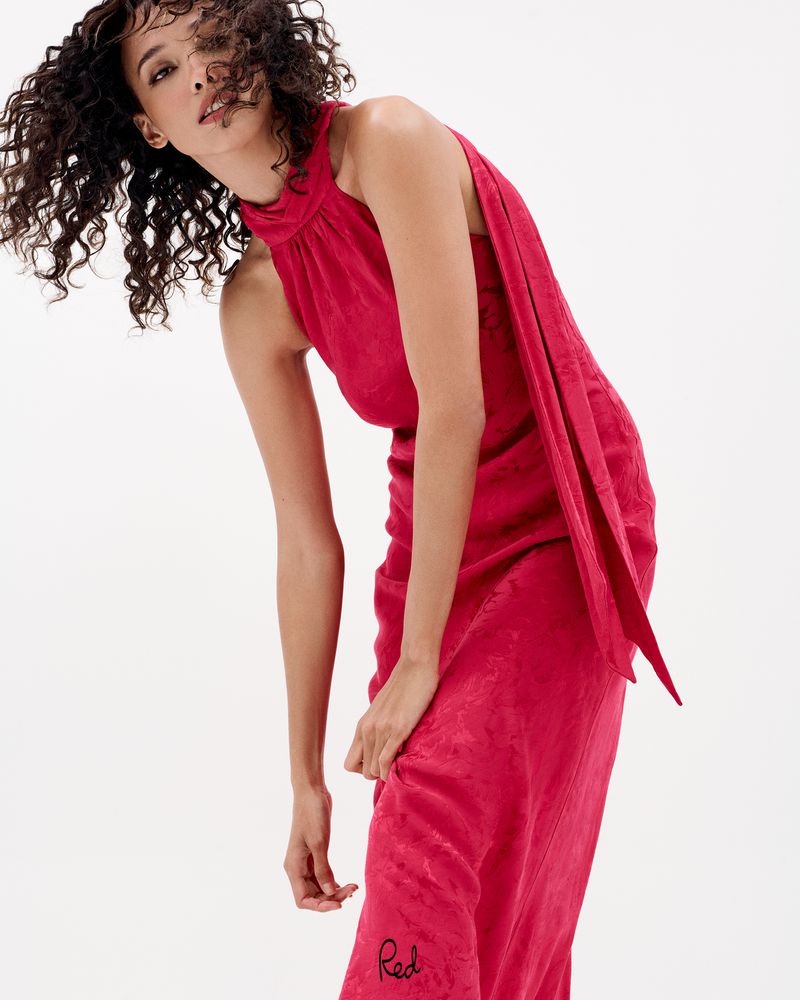 Model Cora Emmanuel posing in a sleeveless high-neck red dress with draped fabric against a clean white seamless backdrop.