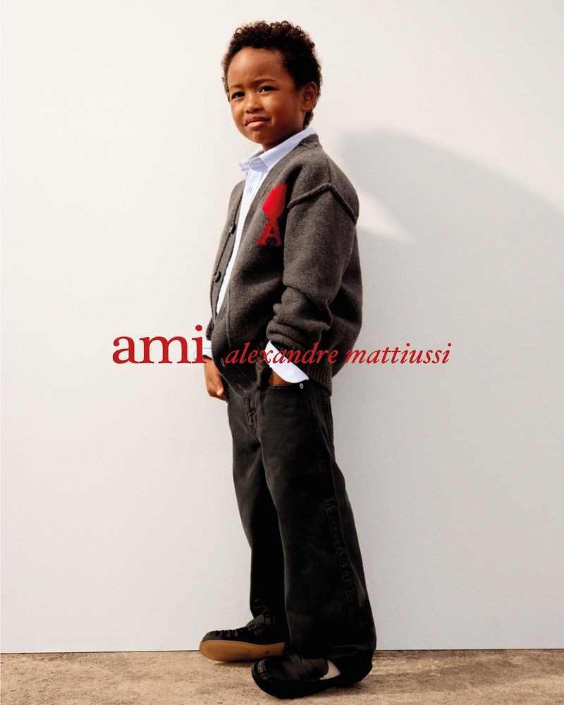 Young boy in a grey knit cardigan and dark denim jeans posing against a white studio wall for Ami Alexandre Mattiussi.
