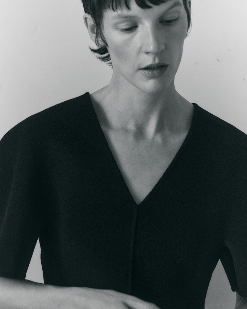 Model Sara Blomqvist wearing a structured black V-neck top against a clean light grey studio backdrop.