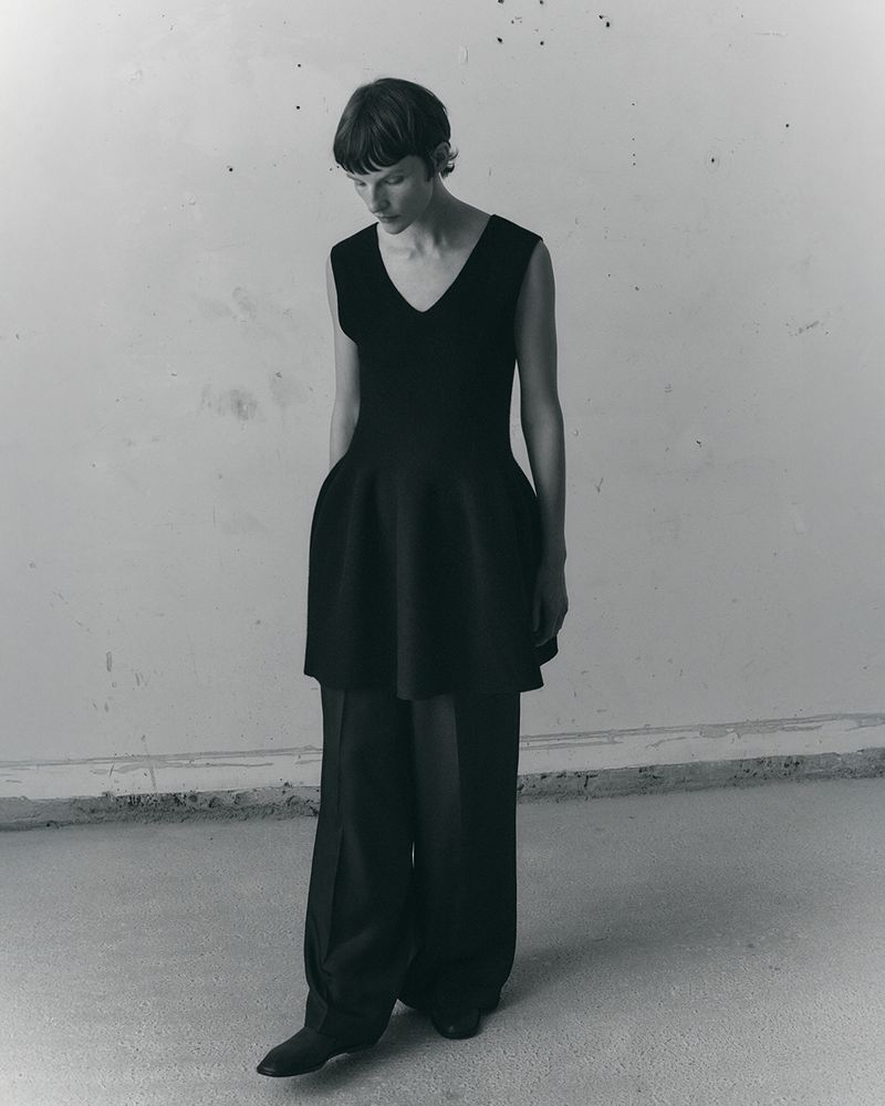 Model Sara Blomqvist wearing a black sleeveless peplum tunic and wide-leg trousers against a textured wall.