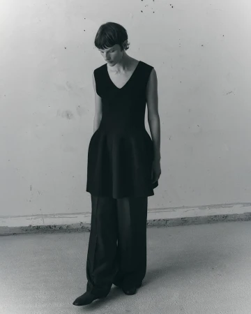 Model Sara Blomqvist wearing a black sleeveless peplum tunic and wide-leg trousers against a textured wall.