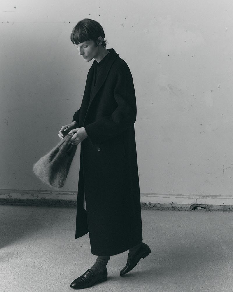 Model Sara Blomqvist wearing a long black tailored coat, dark loafers, and holding a fuzzy tote bag in a minimalist space.