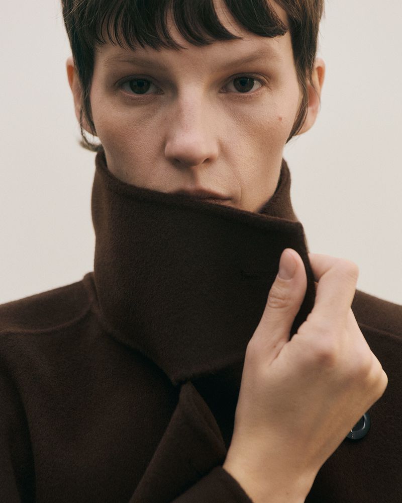 Close-up portrait of a woman with short dark hair, wearing a high-collared dark brown wool coat, pulling the collar up with one hand.