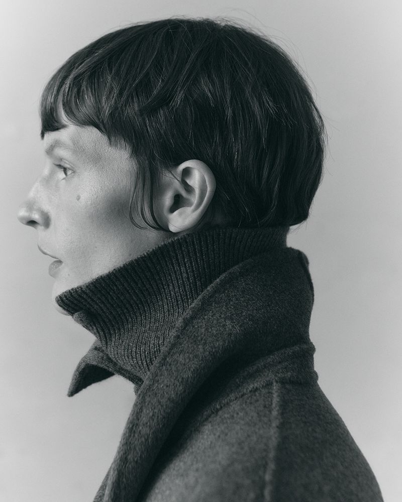 Profile portrait of model Sara Blomqvist wearing a high-collar charcoal knit turtleneck against a muted grey background.