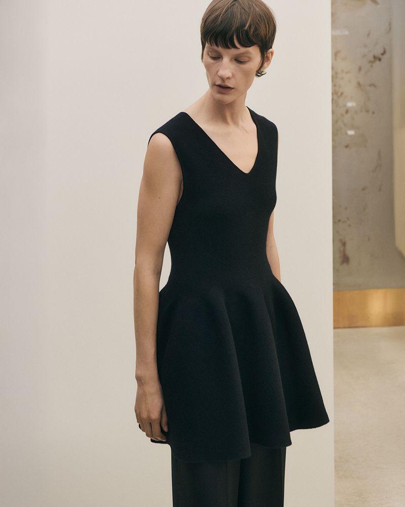 Model Sara Blomqvist wearing a black sleeveless V-neck peplum knit tunic and trousers against a minimalist neutral interior.