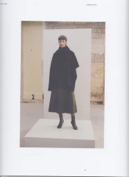 Model Sara Blomqvist in an oversized black knit poncho and olive midi skirt on a white platform for Lisa Yang.