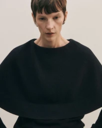 Model Sara Blomqvist wearing a sculptural black knit cape collar sweater against a pale neutral background.