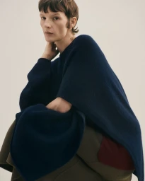 Model Sara Blomqvist wearing a navy knit poncho and olive trousers seated against a plain neutral background.