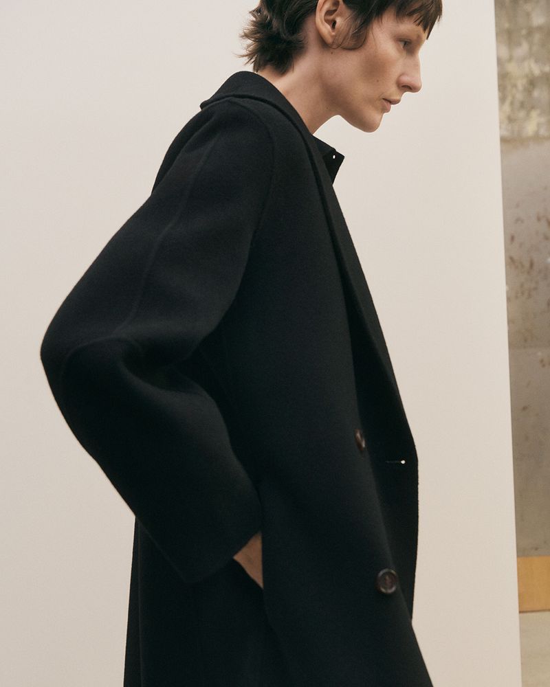 Model Sara Blomqvist captured in profile wearing a structured black overcoat against a neutral minimalist backdrop.