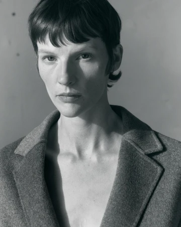 Model Sara Blomqvist wearing a grey tailored wool coat in a close-up portrait against a neutral studio backdrop.