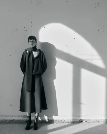 Sara Blomqvist wearing a dark knit sweater, matching scarf, midi skirt, and lace-up boots against a wall with strong shadows.