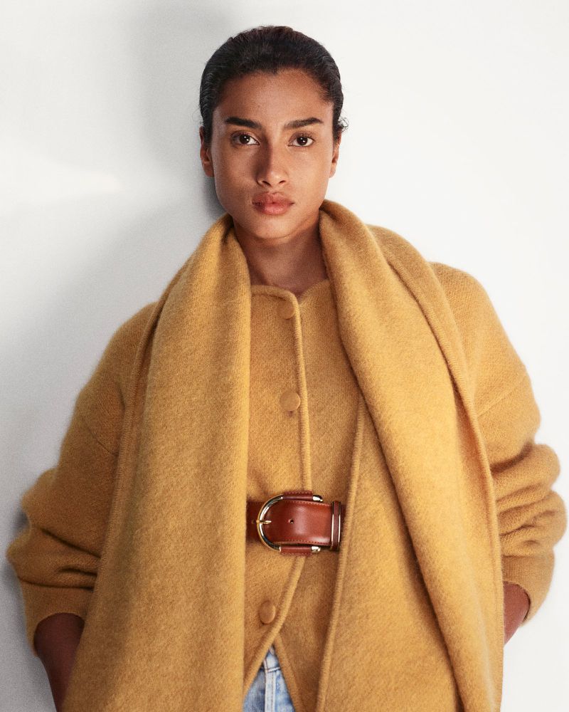 Model Imaan Hammam wearing a mustard yellow wool coat and matching scarf with a brown leather belt against a white backdrop.
