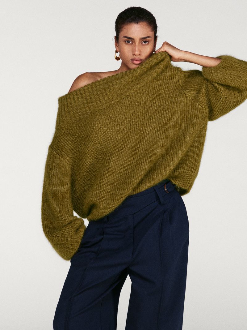 A woman with dark hair wears an oversized olive-green off-the-shoulder mohair sweater paired with navy blue tailored trousers.