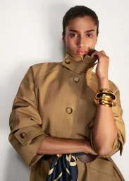 Model Imaan Hammam wearing a high-collar khaki trench coat with a chunky gold cuff bracelet against a studio backdrop.