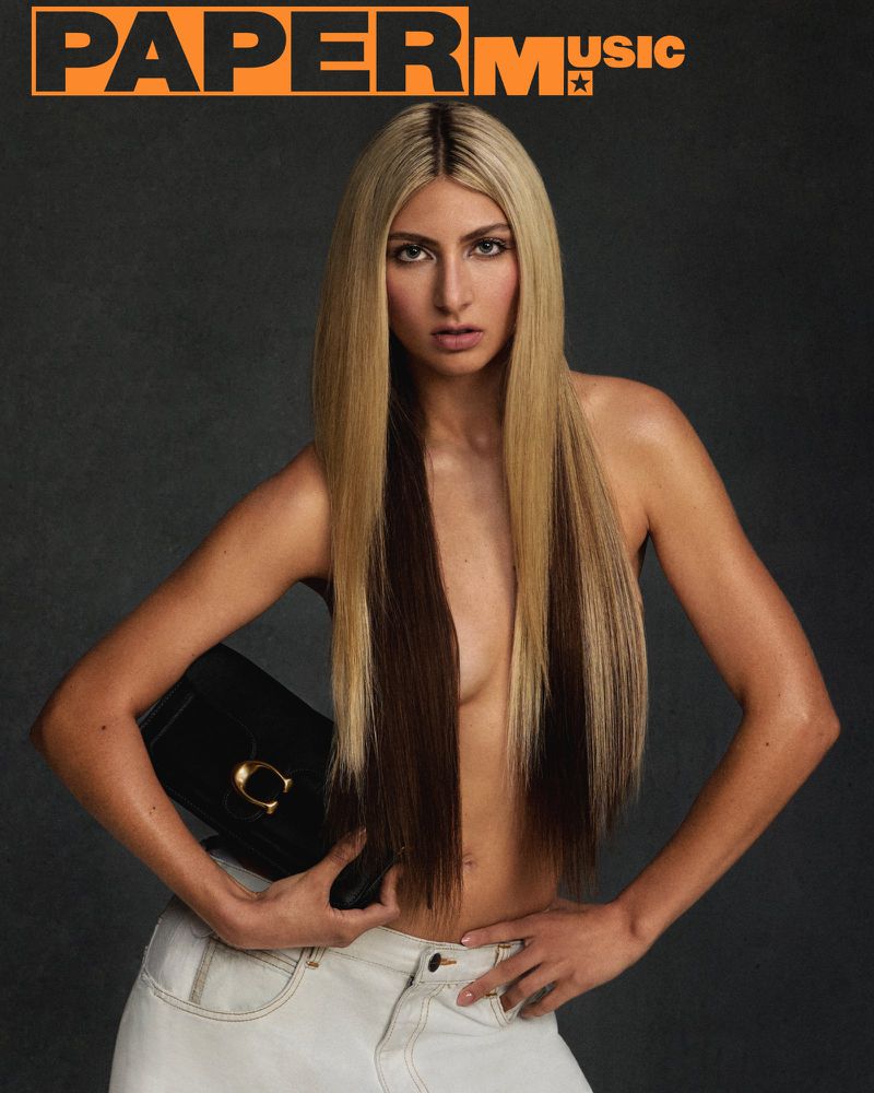 A woman with long blonde and brown hair holds a black clutch bag with a gold clasp against a dark grey background.