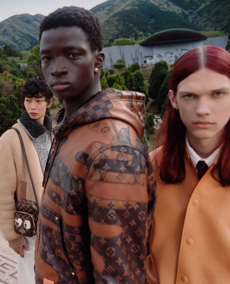 Three fashion models wearing patterned jackets and coats posing outdoors against a backdrop of green hills and architectural structures.