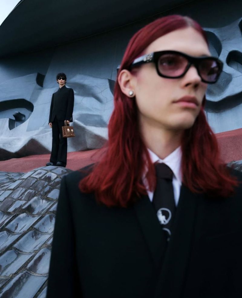 Two people pose in dark suits and sunglasses in front of a brutalist grey concrete building. One holds a briefcase.