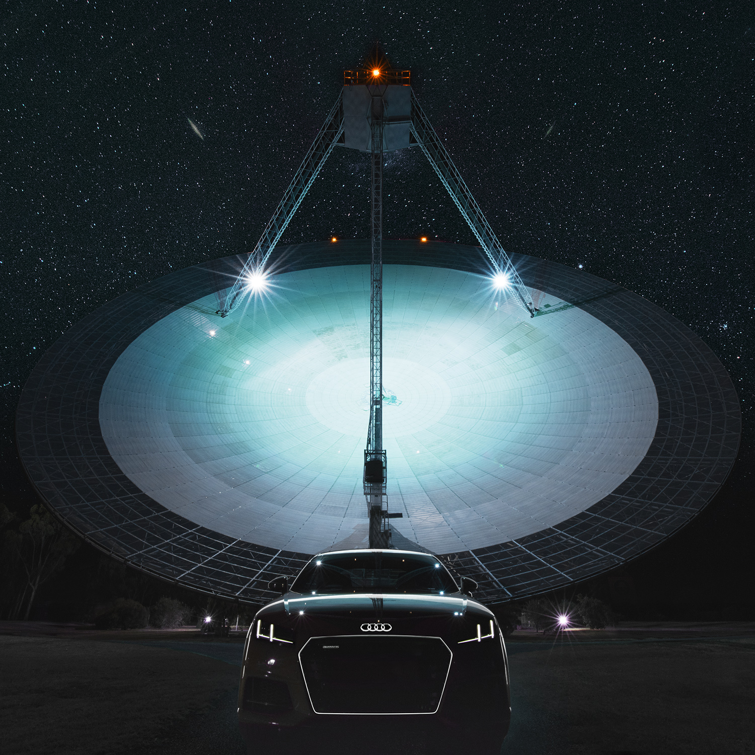 Front view of a black Audi sports car parked at night before a massive illuminated radio telescope dish under a starry sky.