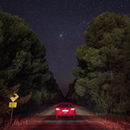 Rear view of a red Audi parked on a dark rural road between dense trees beneath a starry night sky beside a yellow kangaroo road sign.